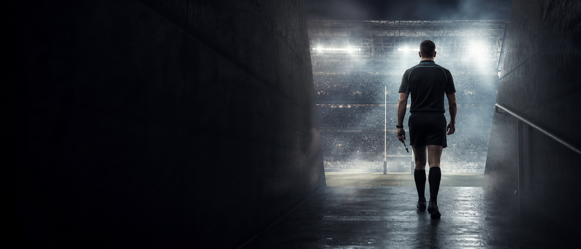 A rugby referee walking through a dark tunnel into a packed floodlit stadium